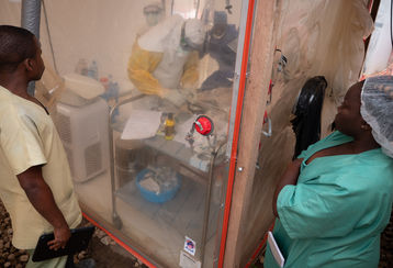 Zwei Frauen in Spitalskleidung stehen vor einem Isolationszelt, während darin ein mit Ebola Infizierter behandelt wird.