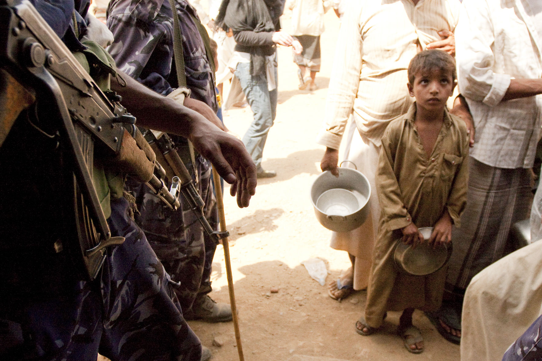 Ein Kind steht in der Schlange bei einer Lebensmittelausgabe. Neben ihm stehen bewaffnete Soldaten.