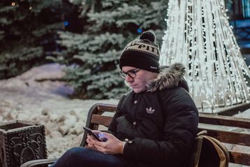 Mann sitzt mit seinem handy auf einer Bank  vor winterlichem Hintergrund